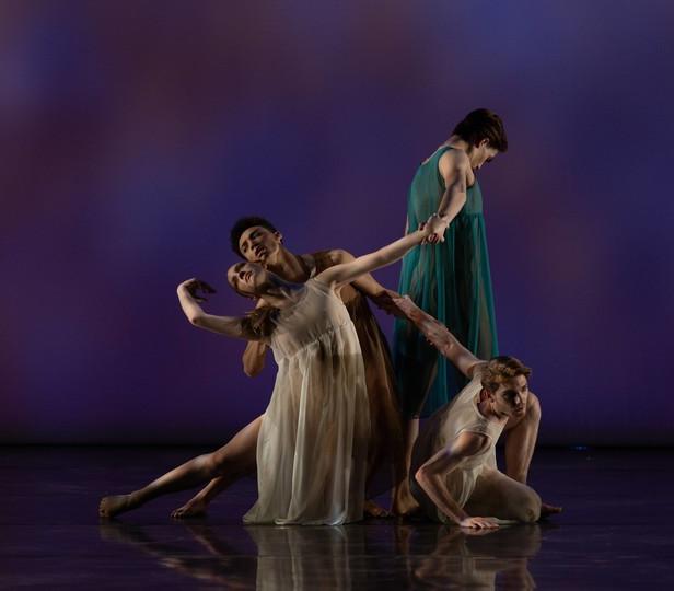 ​​​​​​​Four dancers on stage. One in the foreground is crouched on the floor, propping themselves up with one arm while looking attentively to the side. Behind them, a dancer in a flowing white dress is caught in mid-movement, arms elegantly extended and one leg bent at the knee in a pose that suggests fluid motion. The dancer leans back against another dancer who is standing upright, supporting them with one arm and reaching forward with the other.