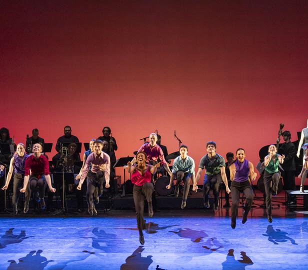 A vibrant stage performance featuring 舞者 energetically leaping into the air. 在后台, a b和 with various musicians is visible, contributing to the dynamic atmosphere of the show. The background is a bold red, enhancing the excitement 和 energy conveyed by the performers. The floor of the stage is adorned with blue patterns, suggesting 运动 和 flow, which complements the dynamic poses of the 舞者.